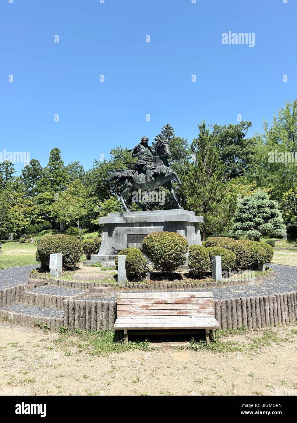 31. August 2025, Takayama, Japan – Bronzemonument eines Samurai-Kriegers zu Pferd mit japanischer Kanji-Inschrift, umgeben von gepflegten Formschnittpflanzen und grünen Bäumen unter einem klaren blauen Himmel. - Smartphone-aufgenommenes Stockfoto
