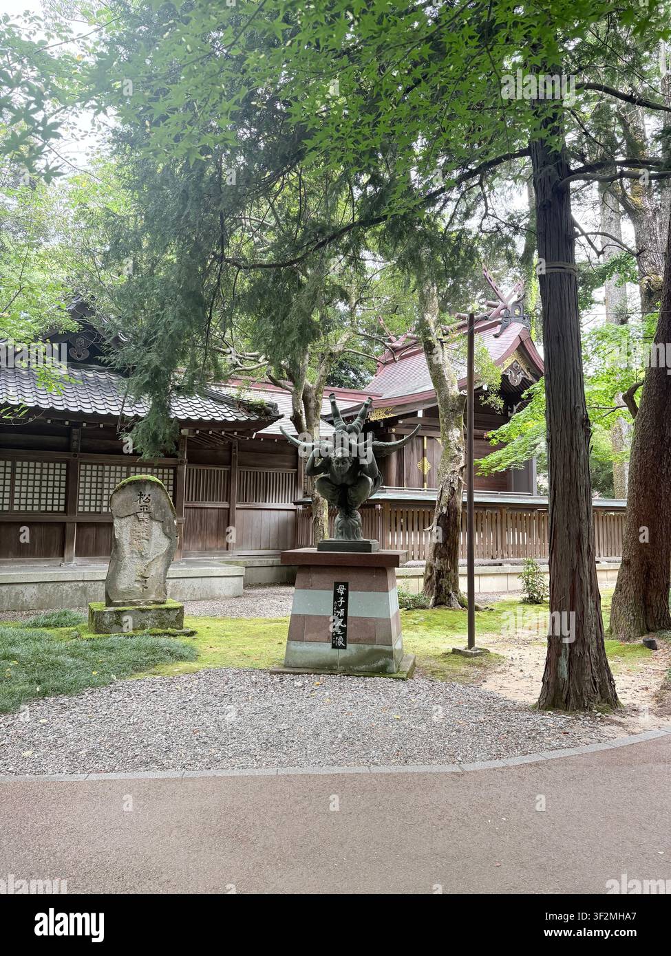 3. September 2025, Kanazawa, Japan – Bronzestatue und Steinmonument am Eingang eines Shintoschreins, umgeben von alten Zedern und traditioneller Holzarchitektur. - Smartphone-aufgenommenes Stockfoto