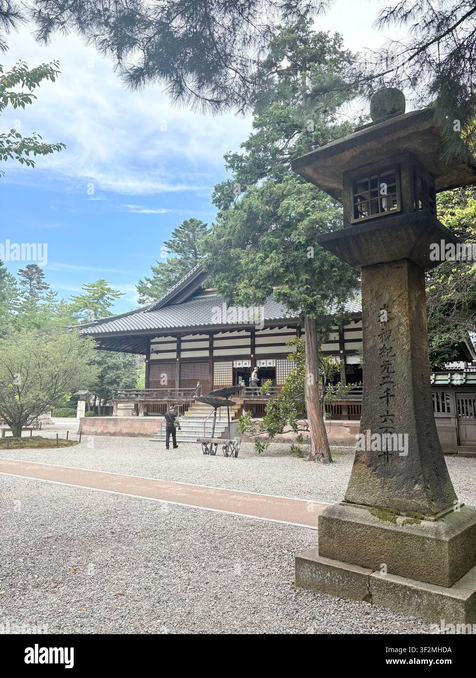 3. September 2025, Kanazawa, Japan – Steinlaterne mit japanischer Kanji-Inschrift am Eingang eines Shintoschreins, traditionelles Holztor und Kiesweg, Kanazawa. - Smartphone-aufgenommenes Stockfoto