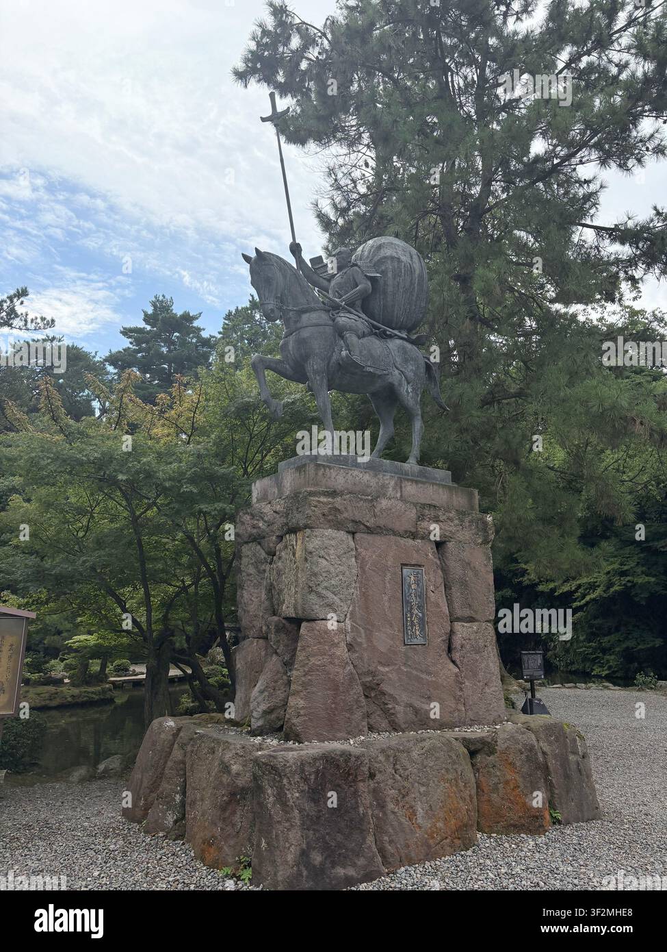 3. September 2025, Kanazawa, Japan – Bronzene Reiterstatue auf steinernem Sockel mit japanischer Inschriftenplakette, umgeben von Kiefern im Park von Schloss Kanazawa. - Smartphone-aufgenommenes Stockfoto
