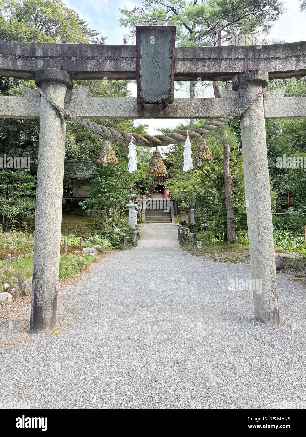3. September 2025, Kanazawa, Japan – Steinerner Torii-Tor mit dickem Shimenawa-Seil und weißen Shide-Bändern an einem traditionellen Shintō-Schrein, üppige Grünpflanzen im Hintergrund. - Smartphone-aufgenommenes Stockfoto