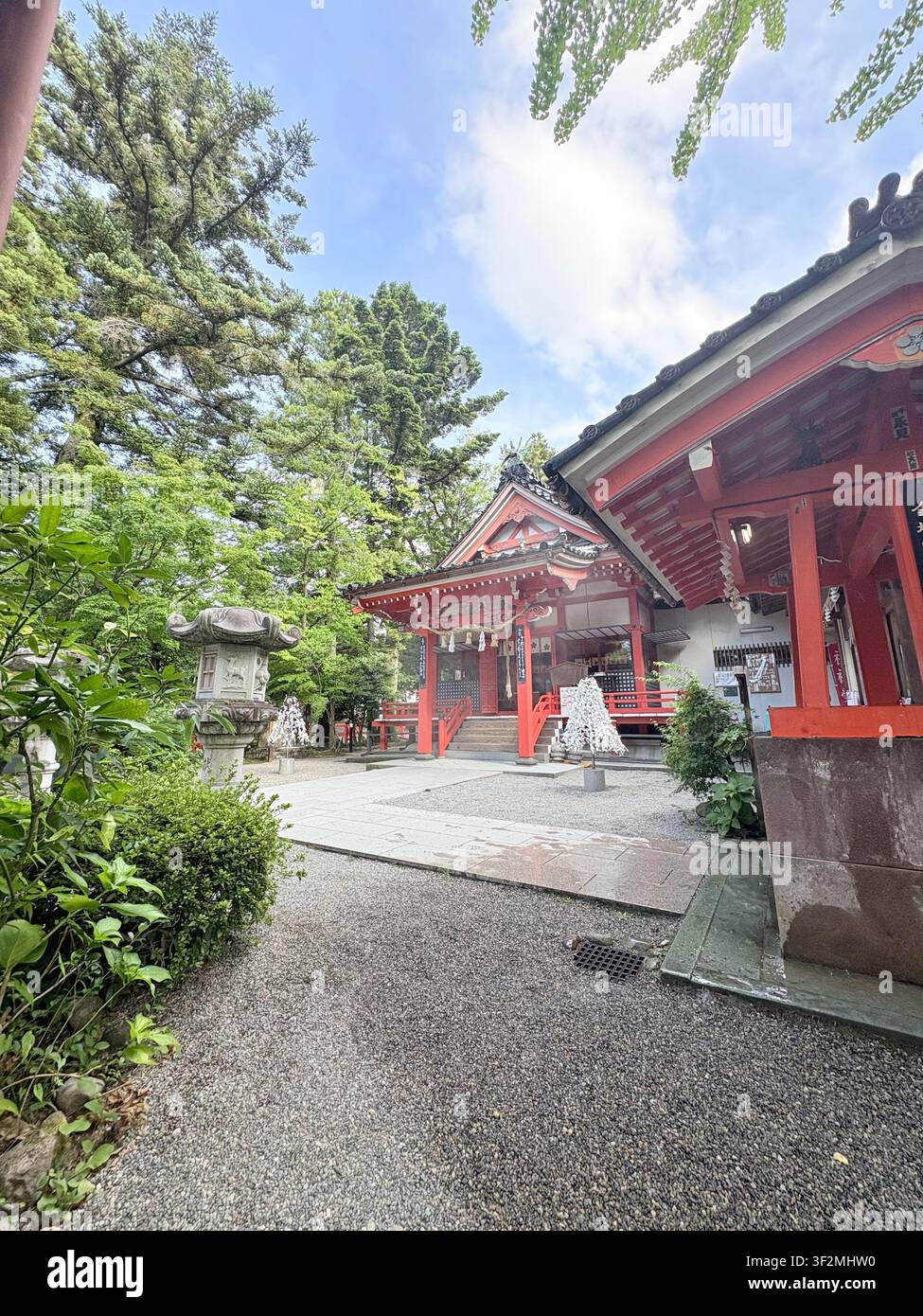 3. September 2025, Japan – Traditioneller rot bemalter Shinto-Schrein mit kunstvollem Pagoden-Dach, Steinlaternen und üppigen grünen Bäumen im Sommerhof. - Smartphone-aufgenommenes Stockfoto