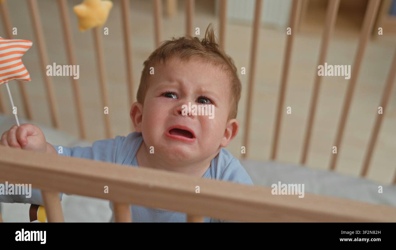 Kleinkind Junge hispanic weint, greift nach orange gestreiftem Sternenspielzeug und packende Gitterstange in hölzernem Kinderbett im Gebäude; Distress braucht Komfort. Stockfoto