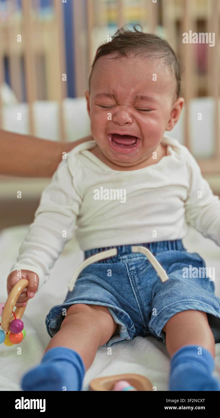 Hispanischer Babyjunge weint, hält eine hölzerne Rassel, während erwachsene Hände Tränen in einem Gebäude-Bettchen abwischen; Trostpflege bei Distress. Stockfoto