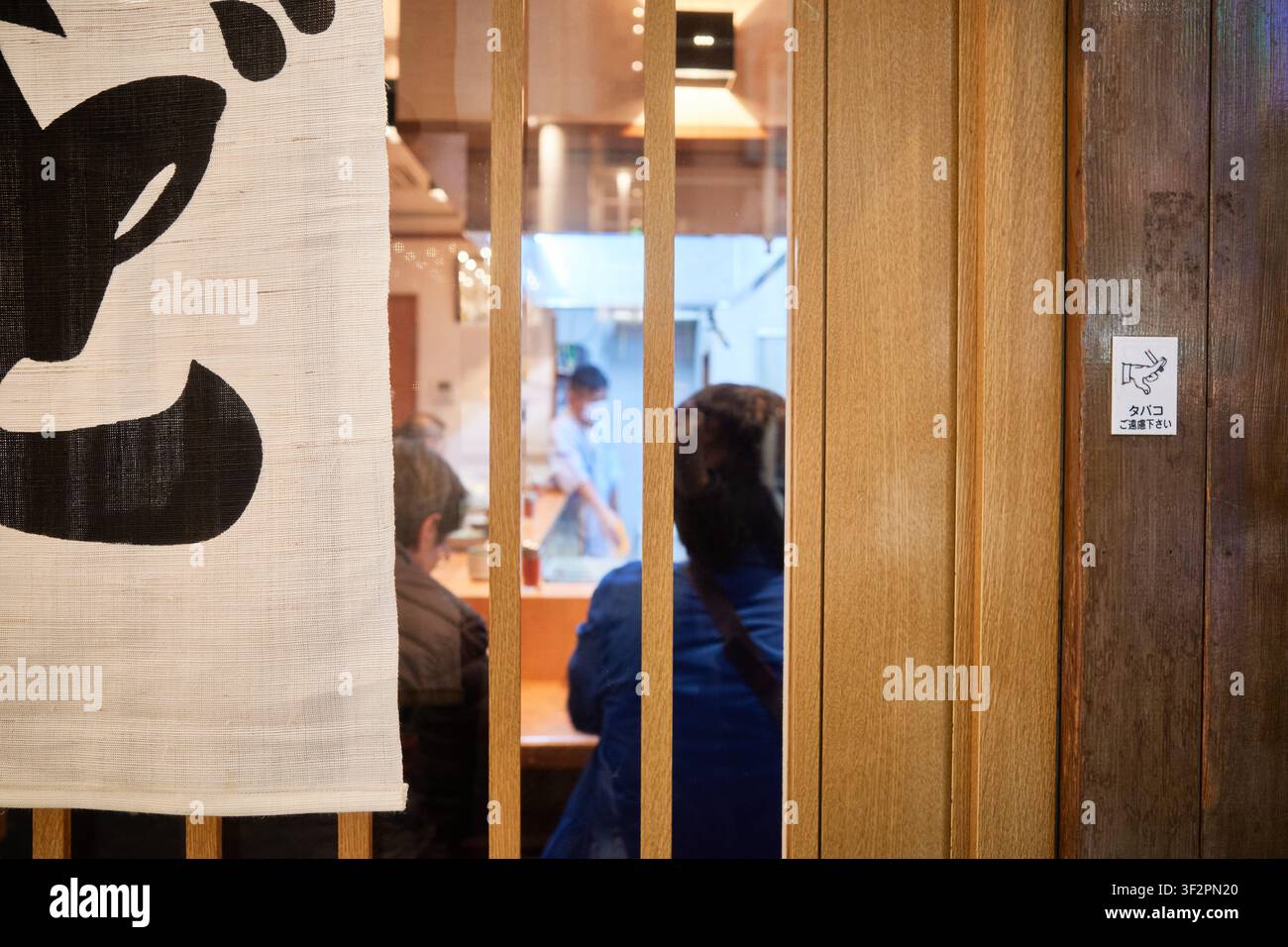 Ein Blick durch holzlattenverschlagene Türen in eine intime Izakaya-Kneipe mit Kunden an einer Theke, perfekt für kulinarische Reiseartikel. Stockfoto
