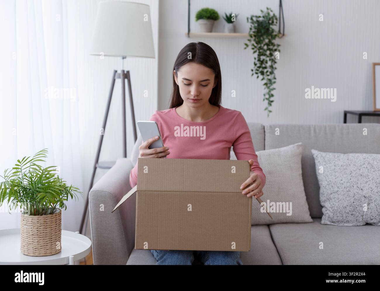 Junge Frau entpackt Bestellung und checkt mit Handy Stockfoto
