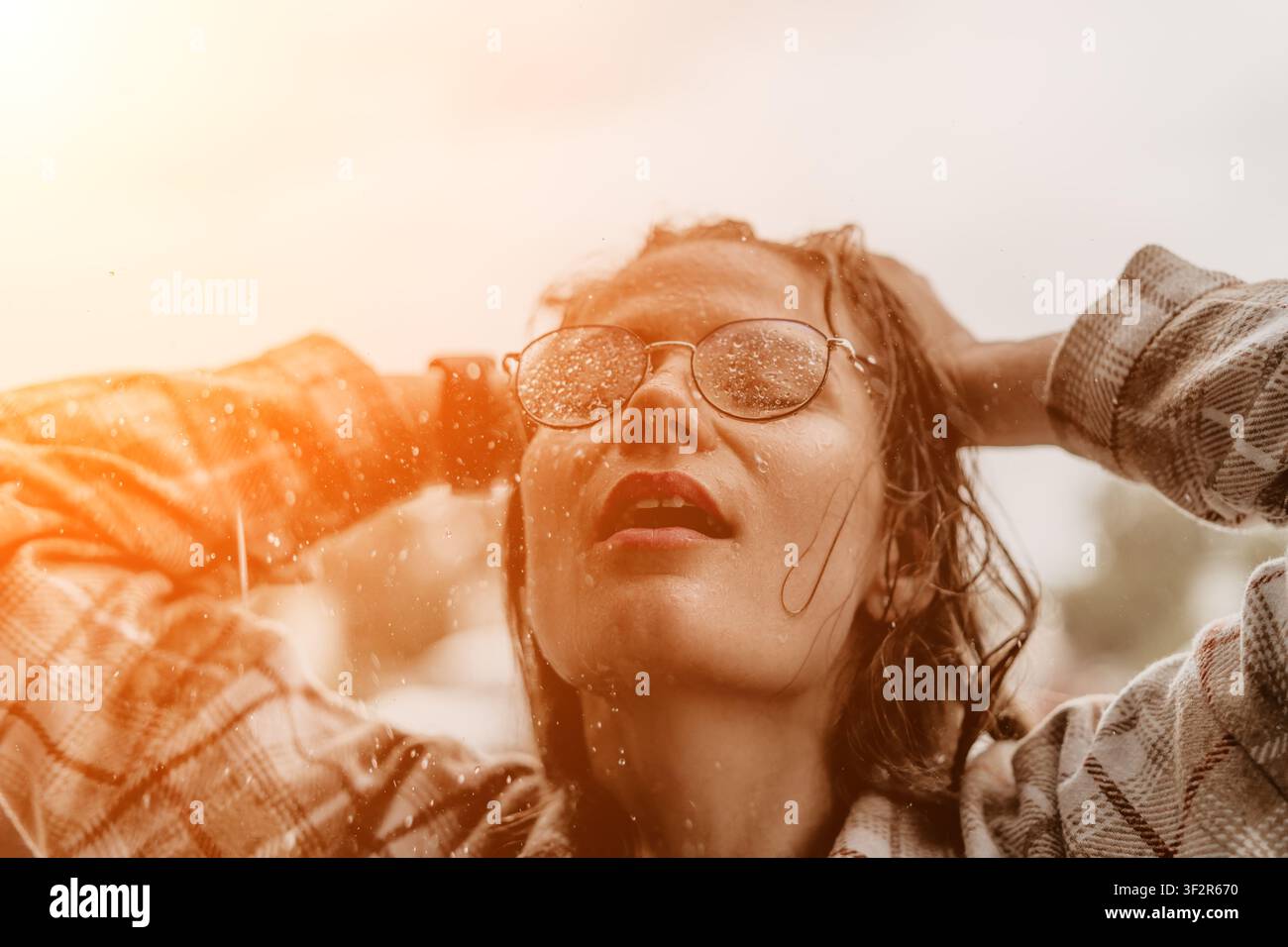 Regenfrau im Freien genießt einen erfrischenden Sommerregen mit zurückgelehntem Kopf, trägt eine Brille und ein kariertes Hemd Stockfoto