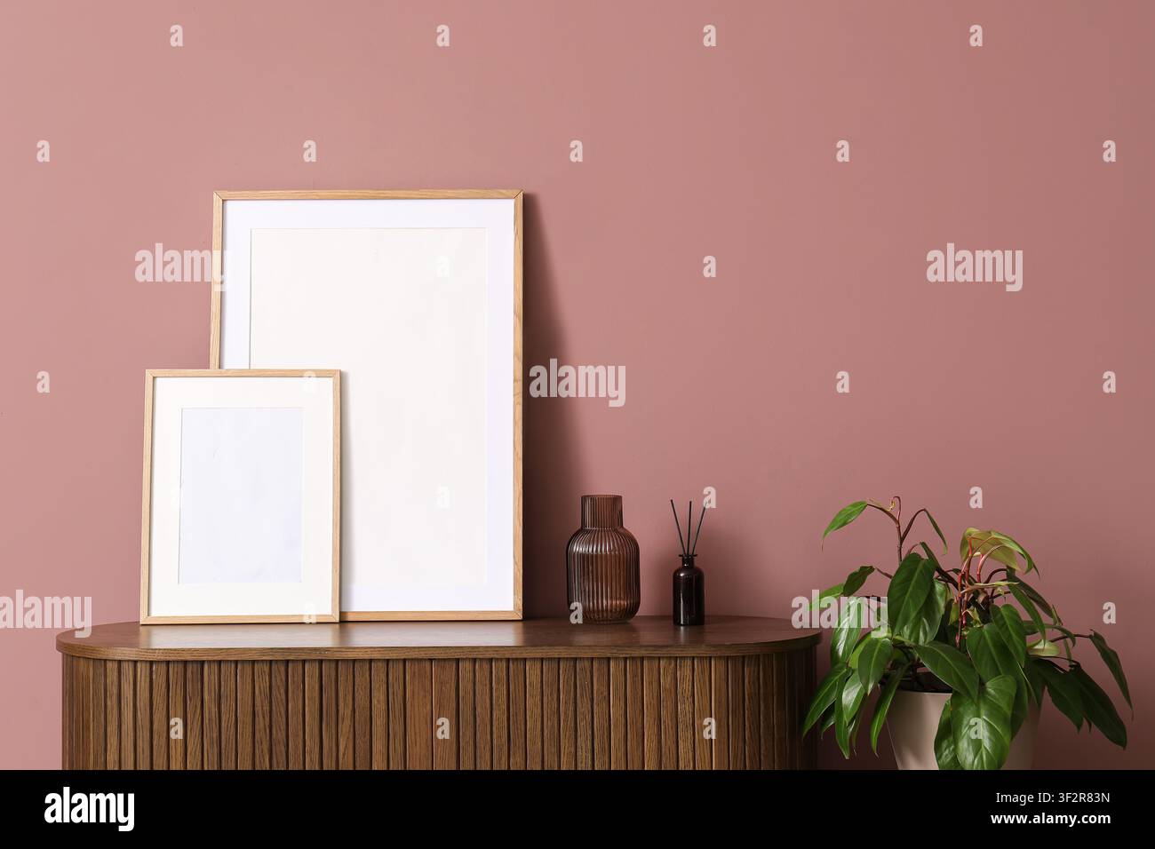 Zwei leere Holzbildrahmen auf einem gerillten Holzschrank mit einer Zimmerpflanze und einem Diffusor vor einer rosa Wand, die Anordnung ist vor einem festen Hintergrund platziert Stockfoto