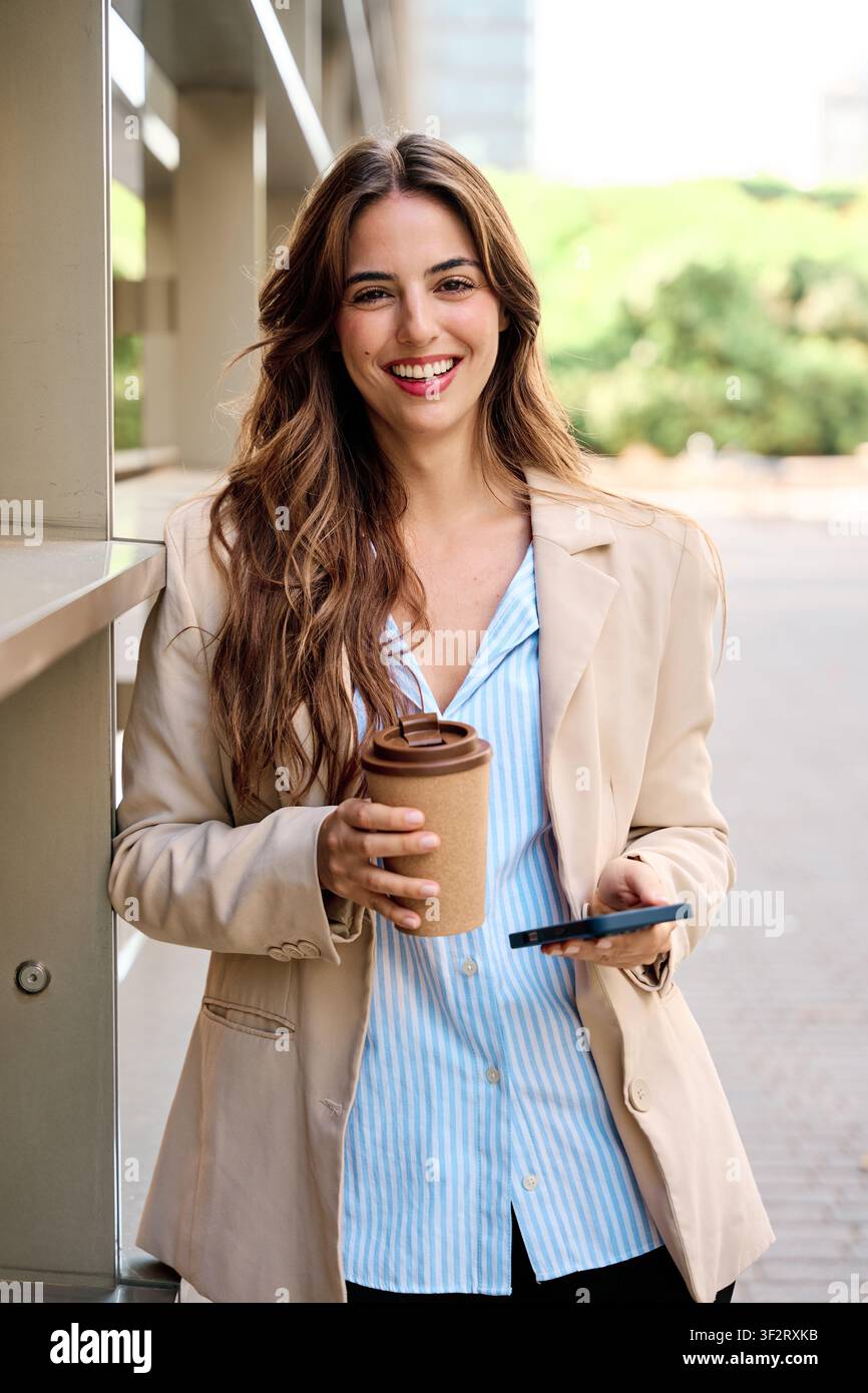 Selbstbewusste junge Geschäftsfrau steht im Freien, hält einen wiederverwendbaren Kaffeebecher und ein Smartphone und präsentiert Professionalität und Erfolg Stockfoto