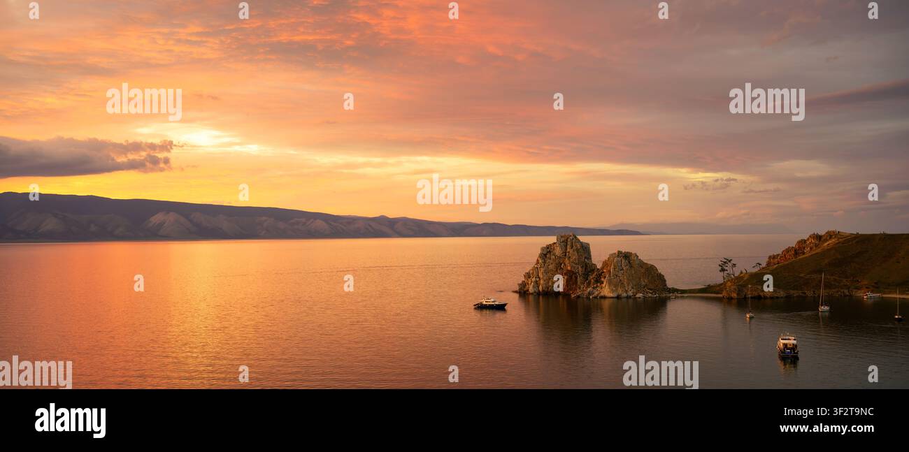 Eine weitläufige Panorama-Landschaft eines lebhaften Sonnenuntergangs über einem ruhigen See. Der Himmel ist erfüllt von sanften orangefarbenen und rosafarbenen Wolken, die sich auf der Wasseroberfläche spiegeln Stockfoto