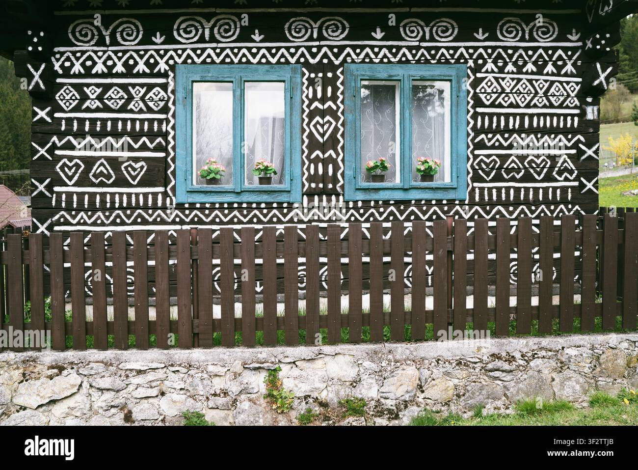 Traditionelle Cicmany-Hausfassade mit ikonischen weißen geometrischen Volksornamentmustern auf dunkler Holzwand mit blauen Fenstern und Holzzaun, Slowakei Stockfoto