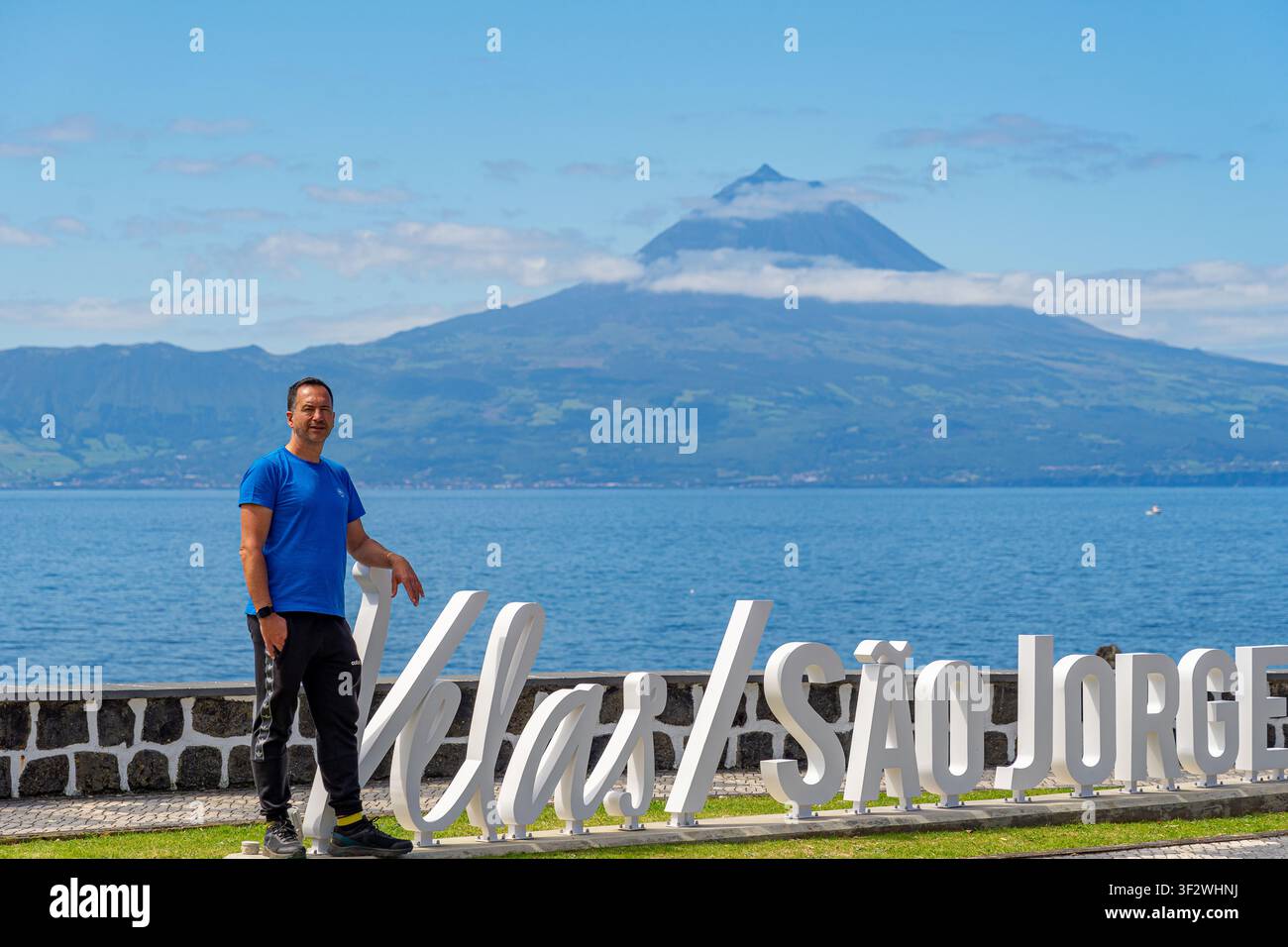 Ein weißer Europäer in einem blauen Hemd steht neben den Buchstaben für die Stadt Velas auf der Insel São Jorge. Ein weißer Europäer in einem blauen Stockfoto