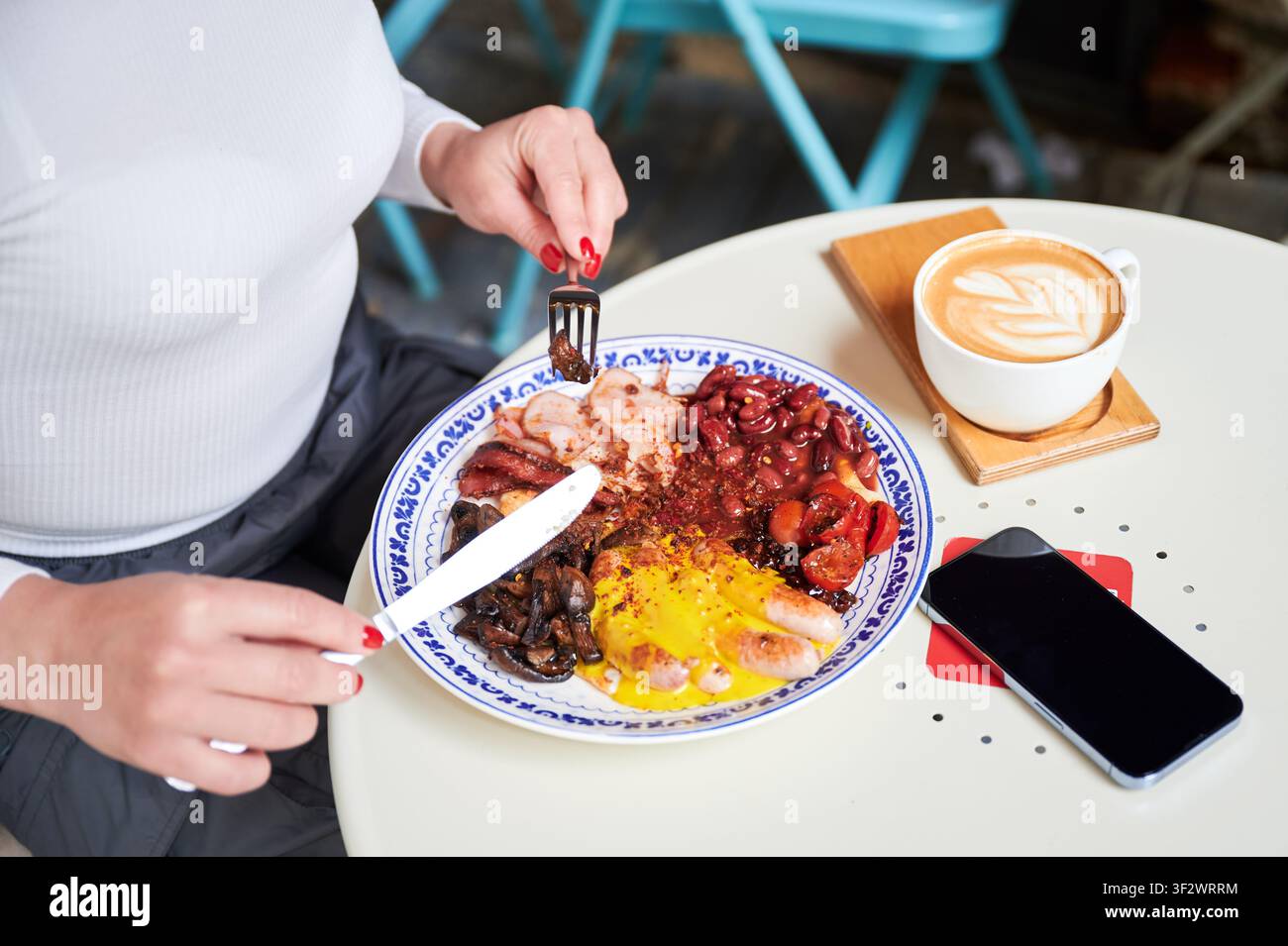 Frau isst herzhaftes Frühstück mit Eiern, Bohnen und Würstchen am Cafétisch, Kaffee und Smartphone in der Nähe, Konzept von urbanem Lebensstil, Comfort Food, Energie und täglicher Routine Stockfoto