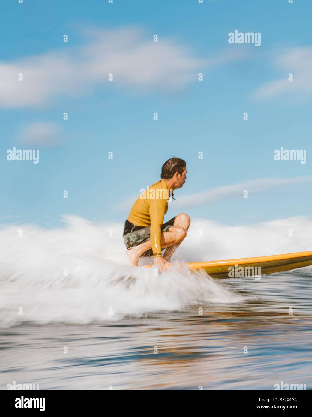 ALA MOANA BOWLS, HONOLULU, HAWAII - 1. APRIL 2022: Ein Mann in einem goldenen Rashguard hockt auf einem orangefarbenen Longboard, während Spritzwasser sich hinter ihm fächert. Stockfoto