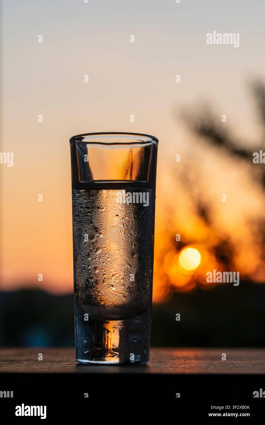 Kaltes Wodka-Schotenglas mit Wassertropfen auf Sonnenuntergang-Bokeh-Hintergrund, Nahaufnahme. Gekühltes, beschlagenes Schotenglas gefüllt mit klarem Alkohol oder Wodka steht da Stockfoto