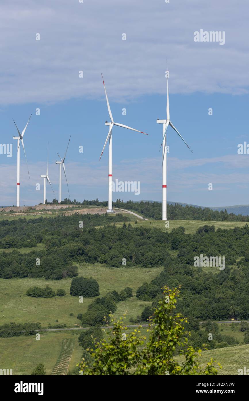Fünf weiße Windturbinen stehen auf einem ländlichen grünen Hügel, der mit Bäumen bedeckt ist, unter einem hellblauen Himmel mit leichten Wolken, was saubere Energieinfrastruktur repräsentiert Stockfoto