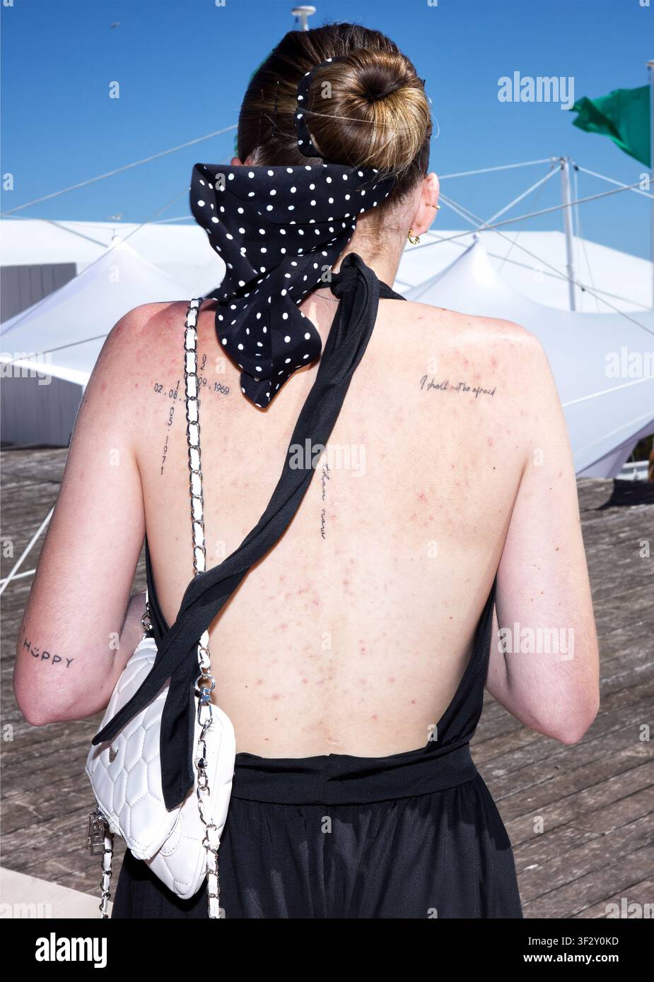 Rückansicht einer jungen erwachsenen Frau, die ein schwarzes rückenfreies Kleid trägt, das verschiedene Tattoos freilegt, mit einem gepunkteten Schal, der um ihr Haar gebunden ist Stockfoto