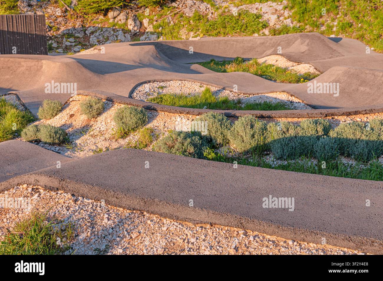 Leere Asphalt-Pumpstrecke mit Böschungen und Hügeln, umgeben von Kies und grünen Büschen, bereit für Radfahren und Outdoor-Sportaktivitäten Stockfoto