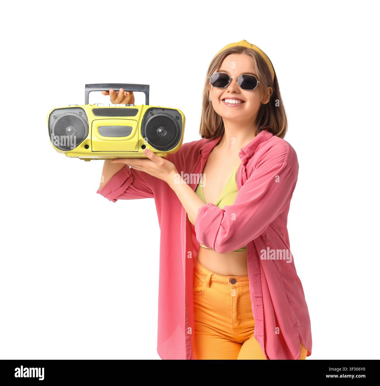 Lächelnde junge Frau mit Sonnenbrille und gelbem Kopfband, die einen Retro-Boombox hält, isoliert auf weißem Hintergrund Stockfoto
