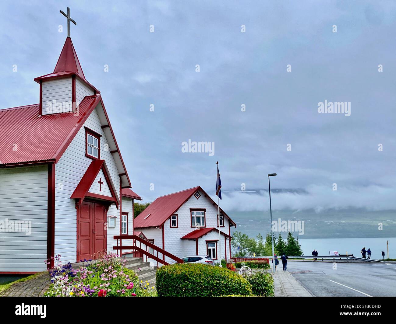 Die Straßenansicht der typischen Kirche mit rotem Dach und weißem Körper in Akureyri City unter einem bewölkten Himmel im Norden Islands - Smartphone-aufgenommenes Stockfoto