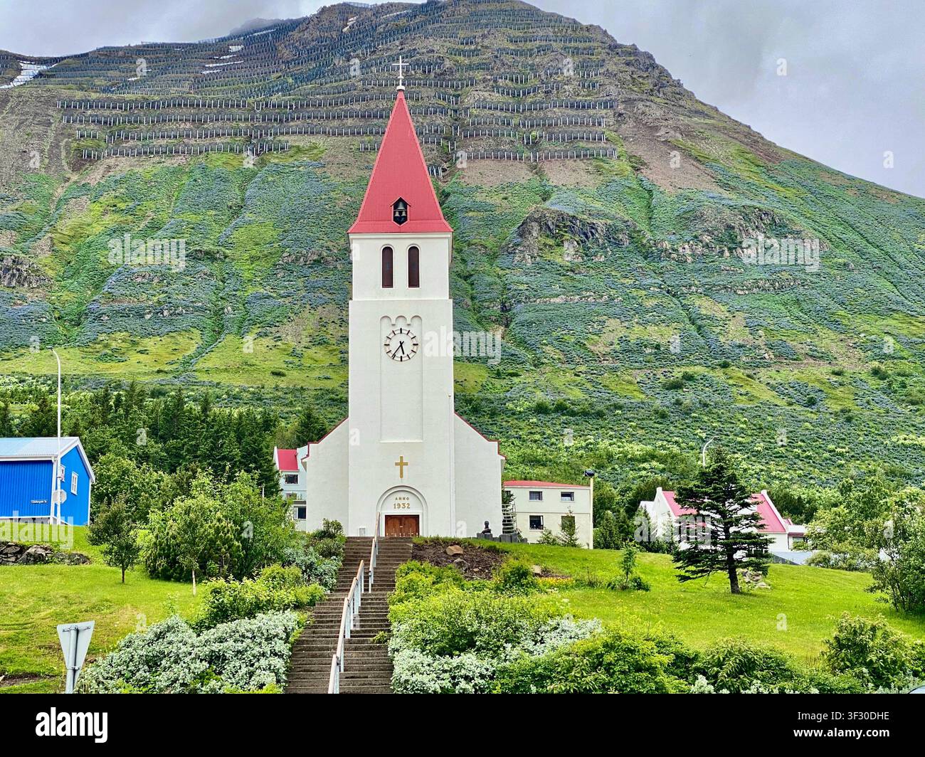 Kirche von Siglufjörður mit Schneebergkette, grünem Gras und Häusern unter einem bewölkten Himmel, Nordisland - Smartphone-aufgenommenes Stockfoto