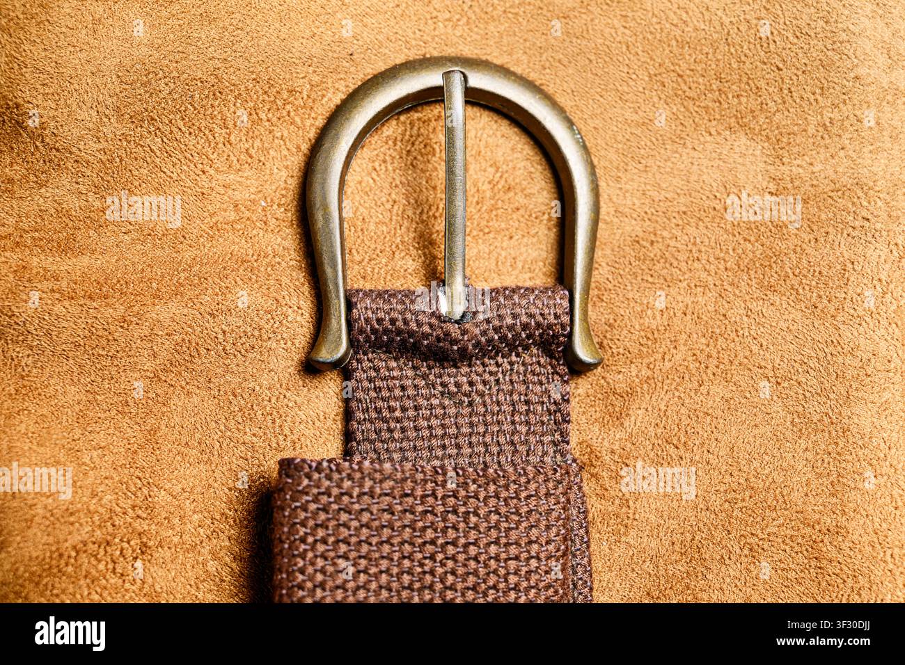 Nahaufnahme einer antiken Messinggürtelschnalle auf einem strukturierten braunen gewebten Riemen, vor einem weichen hellbraunen Wildlederhintergrund. Ideal für Mode und Textur Stockfoto