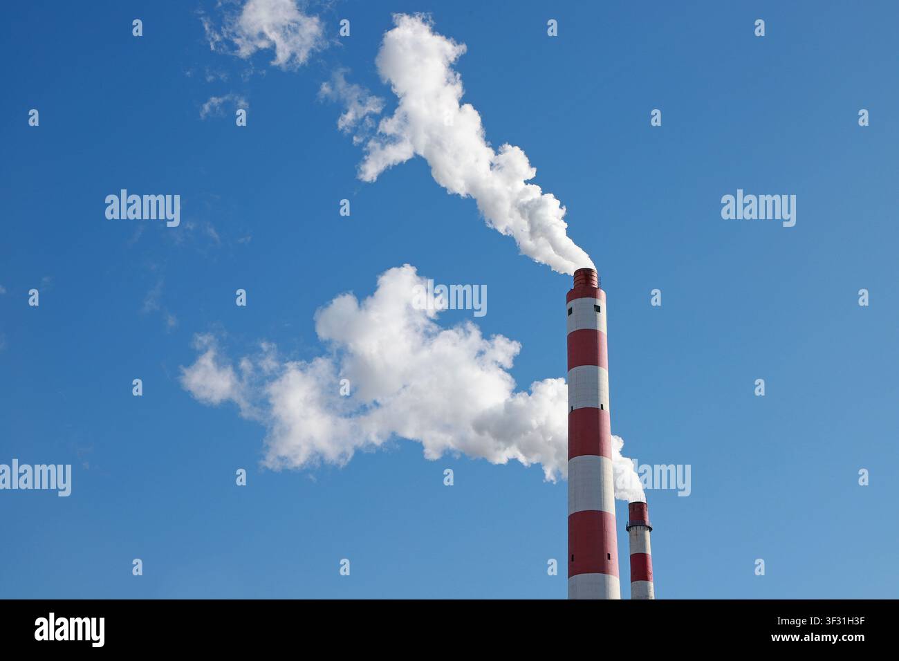 Vor einem klaren blauen Himmel geben zwei rote und weiße Industrie-Schornsteine dicken weißen Rauch ab, der sich mit den natürlichen Wolken vermischt und ein auffälliges I schafft Stockfoto