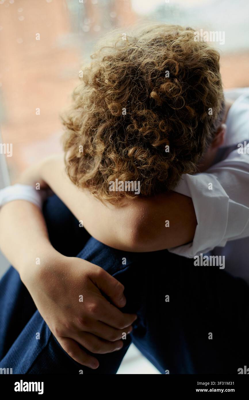 Vertikale Aufnahme eines kaukasischen Jungen, der mit dem Kopf auf den Knien sitzt, sein Gesicht verbirgt und sich in der Nähe eines Fensters zurückzieht, um Traurigkeit oder Stress auszudrücken. Nützlich für Kampagnen zu psychischer Gesundheit, Mobbing, Einsamkeit Stockfoto