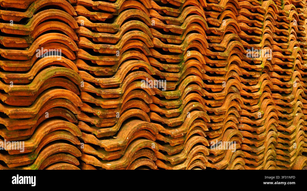 Mehrere Reihen ordentlich gestapelter Terracotta-Keramikdachziegel bilden einen auffälligen orange-grünen Welltexturhintergrund für Dachkonzepte. Stockfoto