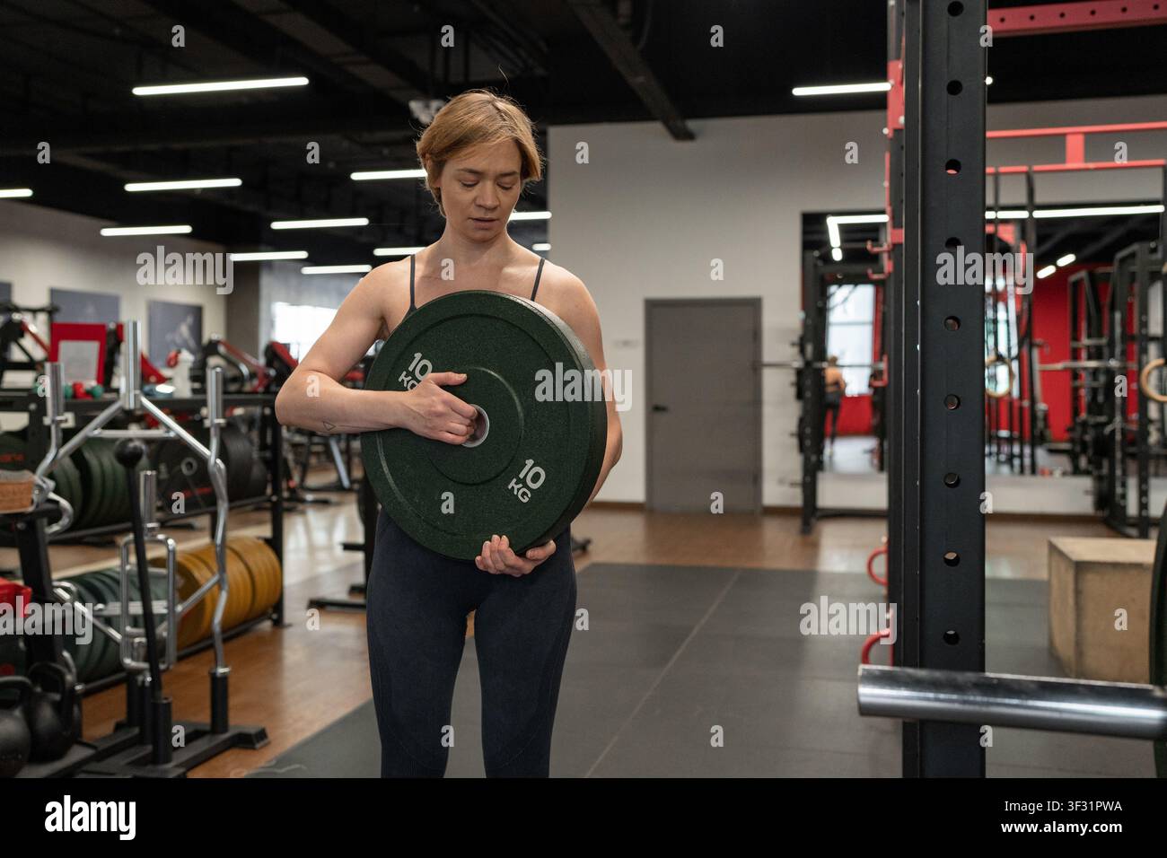 Feminine Athletin lädt schwere Gewichtsplatte auf die Stange in der Hantelbank in einem hellen modernen Fitnessstudio, konzentrierter Ausdruck, starke Haltung und sichtbare Anstrengung Stockfoto