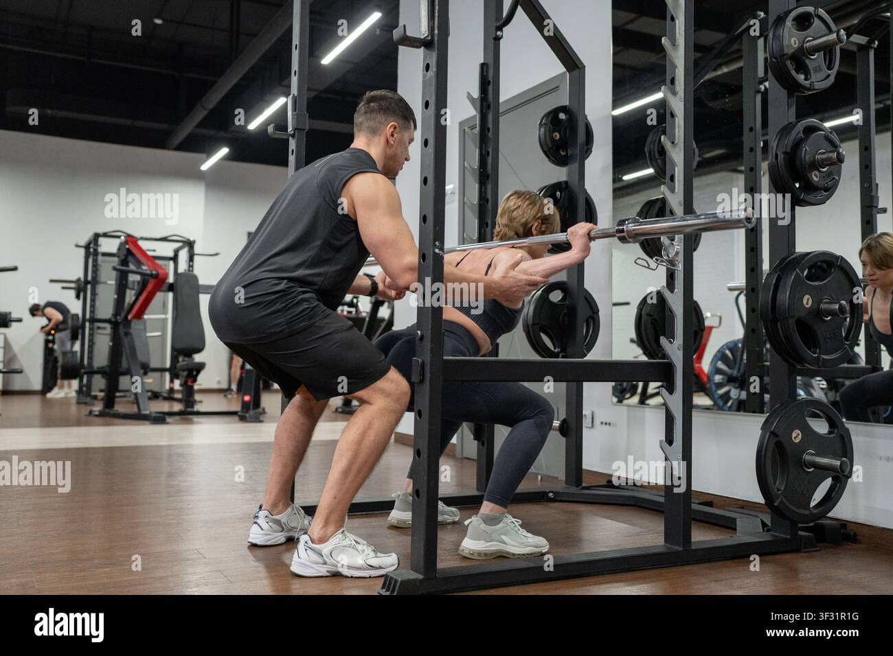 Männlicher Trainer unterstützt weibliche Athletin beim Back-Squat mit der Hantelstange in einem modernen Fitnessstudio mit Rack, Gewichten und konzentrierter Trainingsunterstützung sichtbar im gesamten Raum Stockfoto