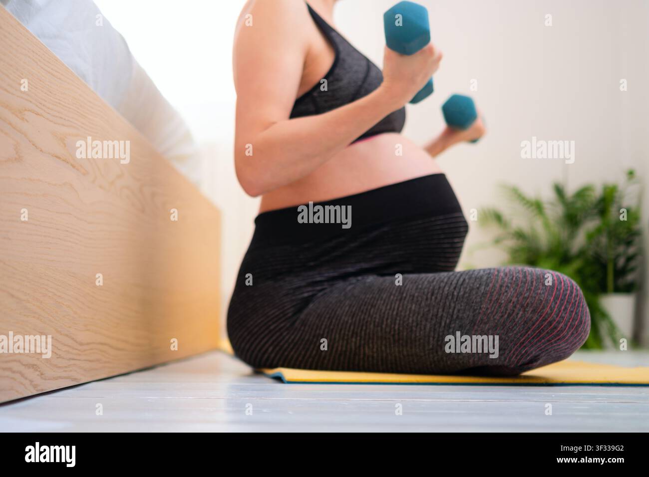 Eine schwangere Frau sitzt auf einer Yogamatte und hält eine Hantel, sie praktiziert Pränatalyoga auf dem Boden zu Hause. Die Mutter und Sportlerin zeigt ruhiges, achtsames Wohlbefinden und friedliche, ausgewogene Fitness in einem Heimfitnessstudio. Stockfoto