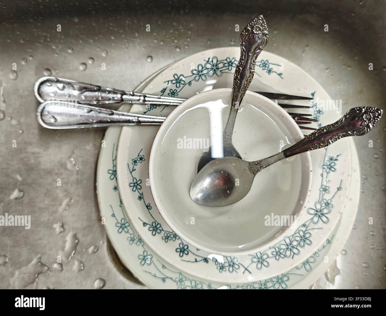 Geschirr und Besteck ruhen in einem Spülbecken, das mit Wassertropfen gefüllt ist, nach einer Mahlzeit. Teller, Schüsseln und Utensilien sind ordentlich angeordnet. Stockfoto