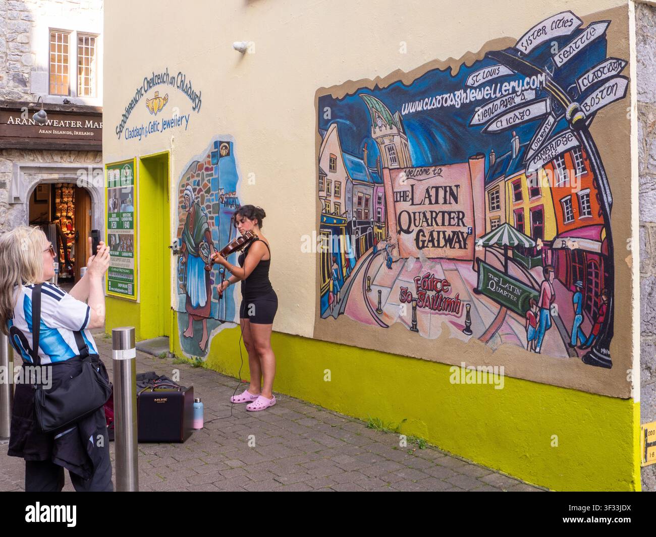 Am Eingang zum Lateinviertel in der Stadt Galway unterhält ein straßmusizierender Geiger die Besucher vor einem Wandgemälde, während ein Tourist ein Foto macht Stockfoto