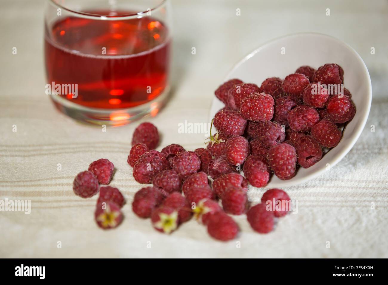 Eine horizontale Nahaufnahme von reifen roten Himbeeren, die aus einer weißen Keramikschale auf ein Leinentischtuch fallen. Ein Glas roter Saft ist sanft unscharf Stockfoto
