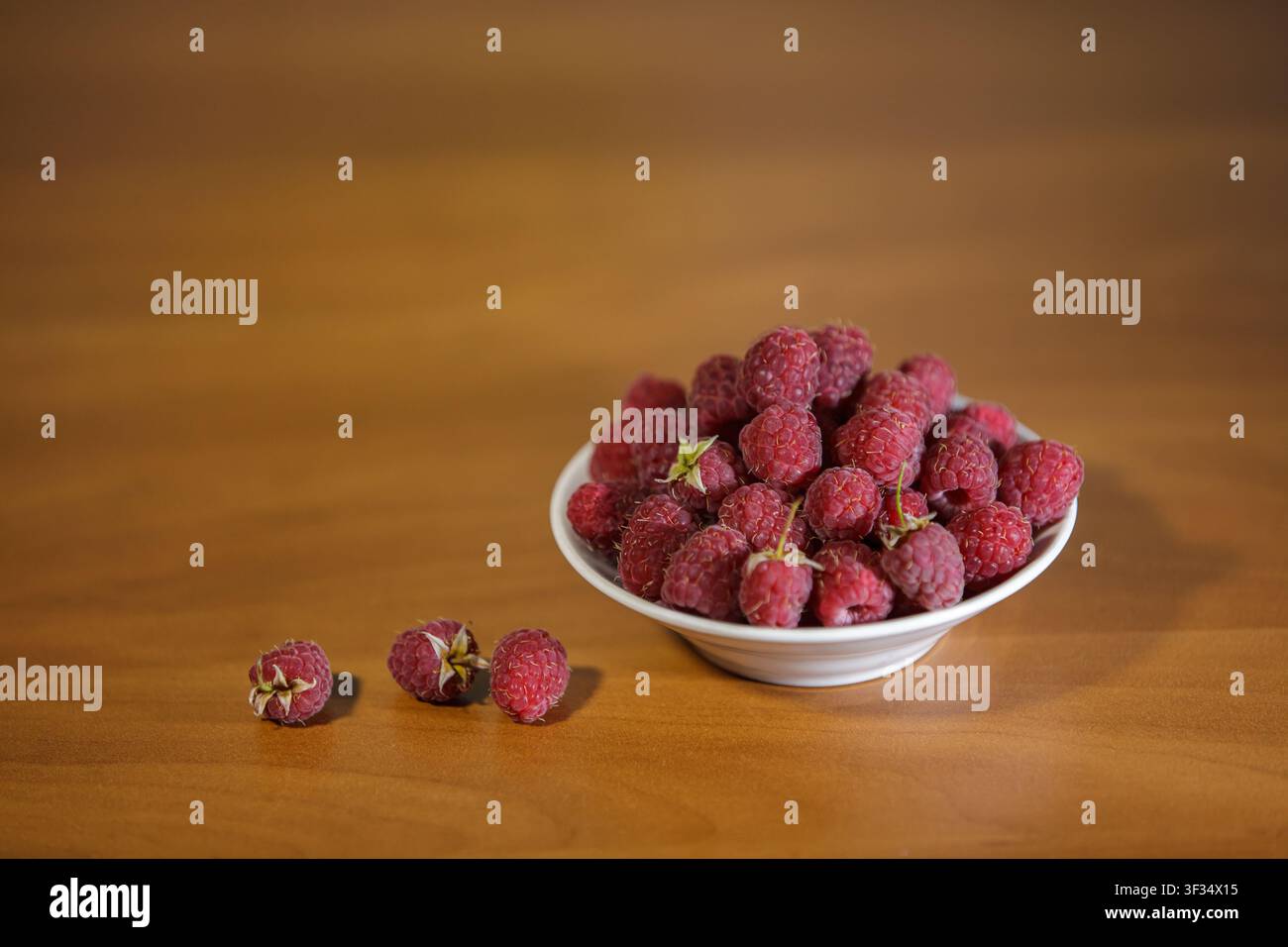 Eine Nahaufnahme-Horizontalaufnahme einer kleinen weißen Keramikschale, die bis zum Rand mit frischen roten Himbeeren gefüllt ist. Drei einzelne Beeren liegen auf dem smoo Stockfoto