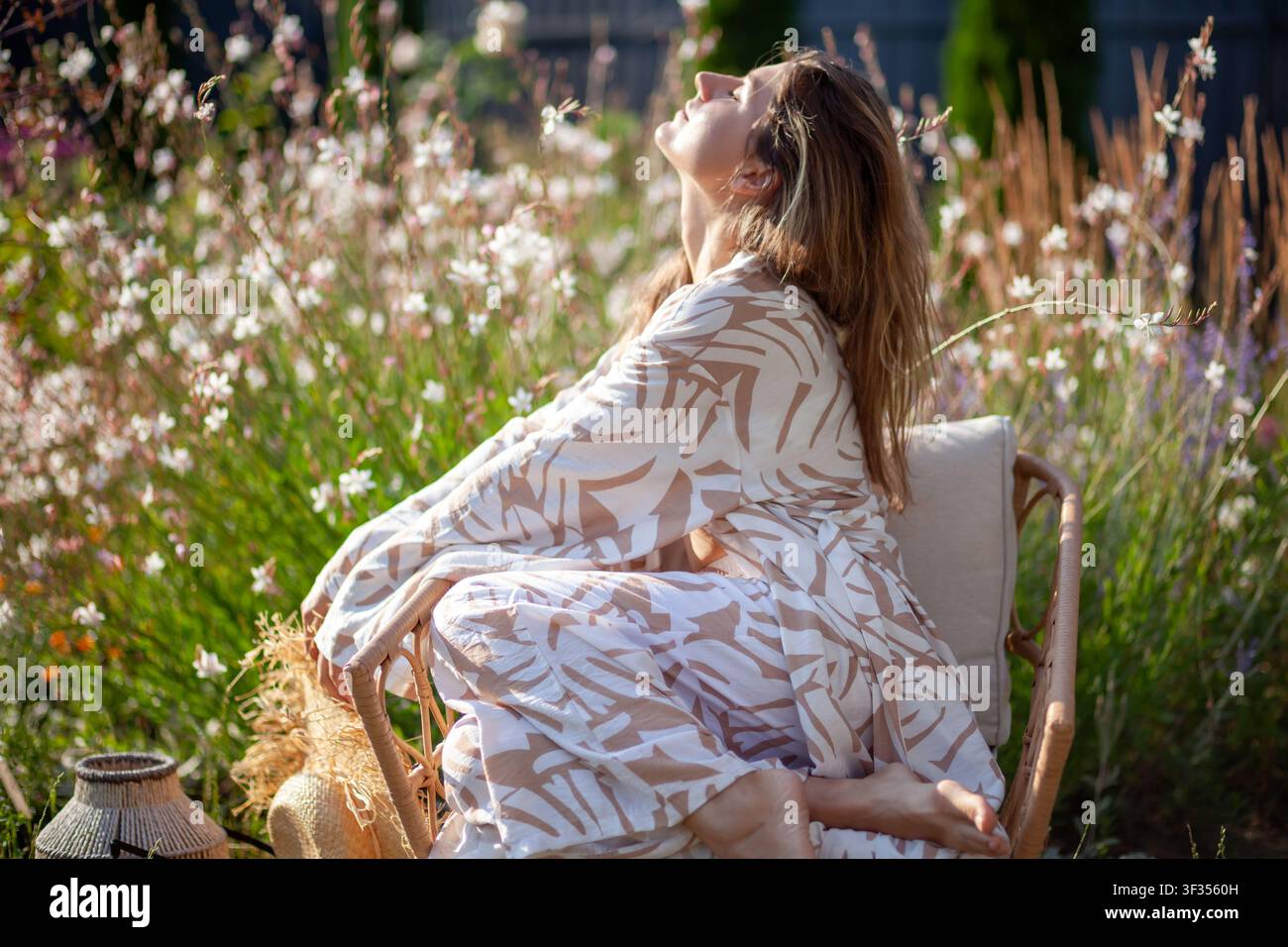 Junge Frau entspannt sich auf einem Stuhl im Sommergarten, trägt ein stilvolles Gewand und hält einen Strohhut. Die Dame atmet, genießt die Sonne zwischen Blumen Stockfoto