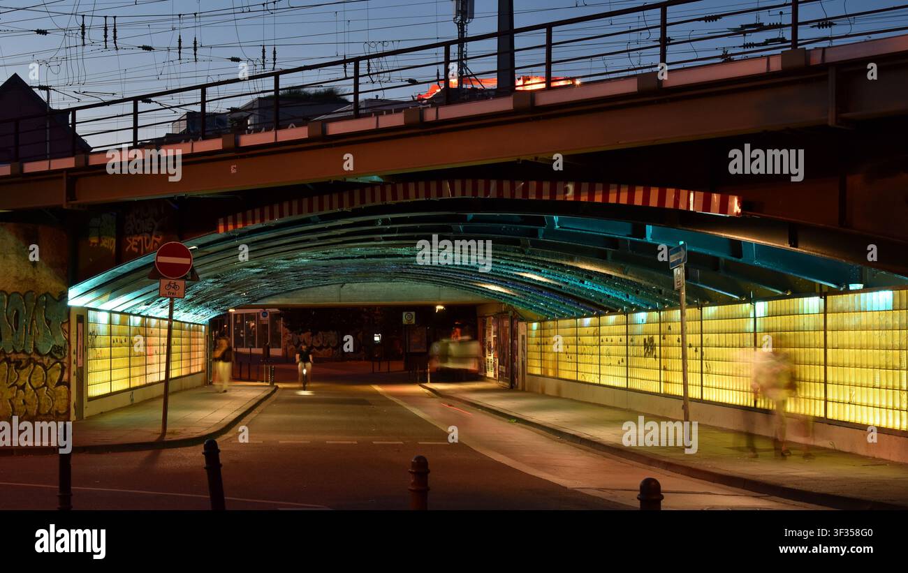 Geisterhafte Bilder von Fußgängern und Radfahrern, die die beleuchtete Marzellenstrasse passieren, während sie unter einer Eisenbahnbrücke in Köln, Deutschland, verlaufen. Stockfoto