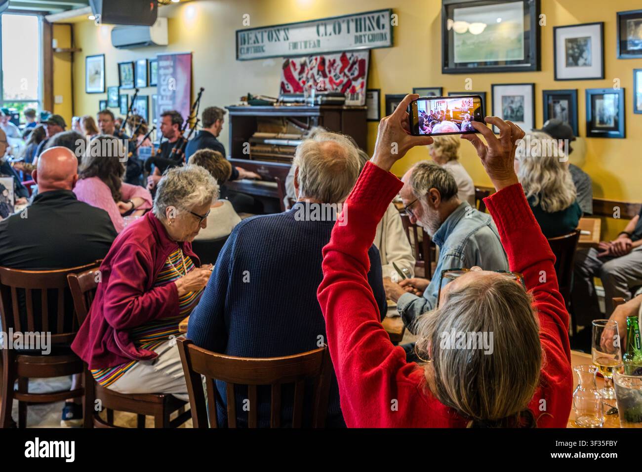 Eine Besucherin filmt spielende Dudelsackspieler auf ihrem Smartphone im Red Shoe Pub in Mabou, Nova Scotia, Kanada. An der Wand hängt das historische Schild ‚Beaton’s Clothing‘, eine Erinnerung an die frühere Nutzung des Gebäudes als Gemischtwarenladen. Mabou, Nova Scotia, Kanada Stockfoto