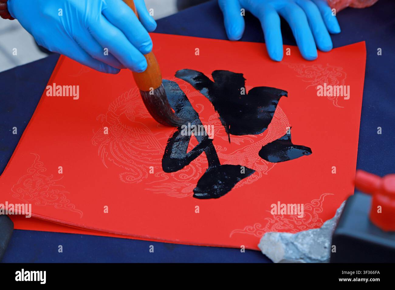 Nahaufnahme einer traditionellen chinesischen Kalligrafie, die auf rotem Xuan-Papier geschrieben wird. Eine Hand, die blaue Schutzhandschuhe trägt, hält einen schwarzen Tintenpinsel, creat Stockfoto