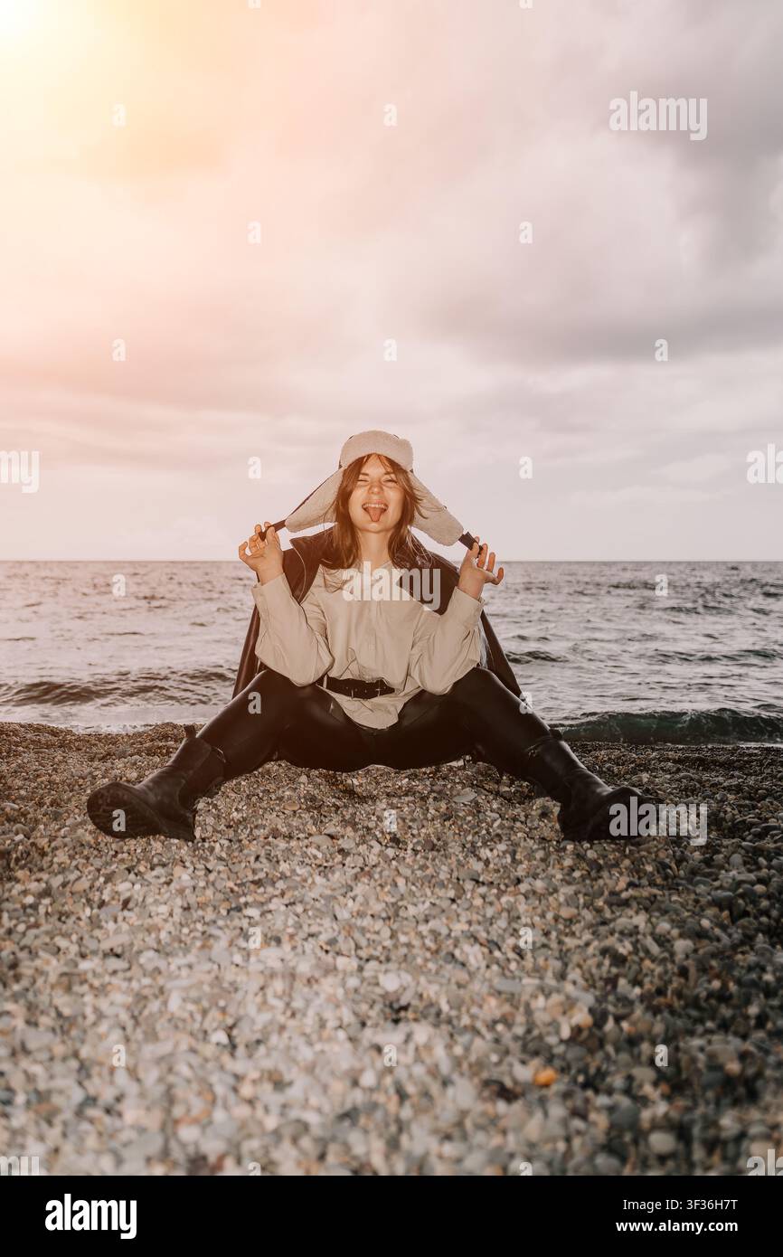 Frau am Strand mit Uschanka, verspielte Frau sitzt auf einem Kiesstrand, trägt einen Winterhut und streckt die Zunge heraus an einem bewölkten Tag Stockfoto