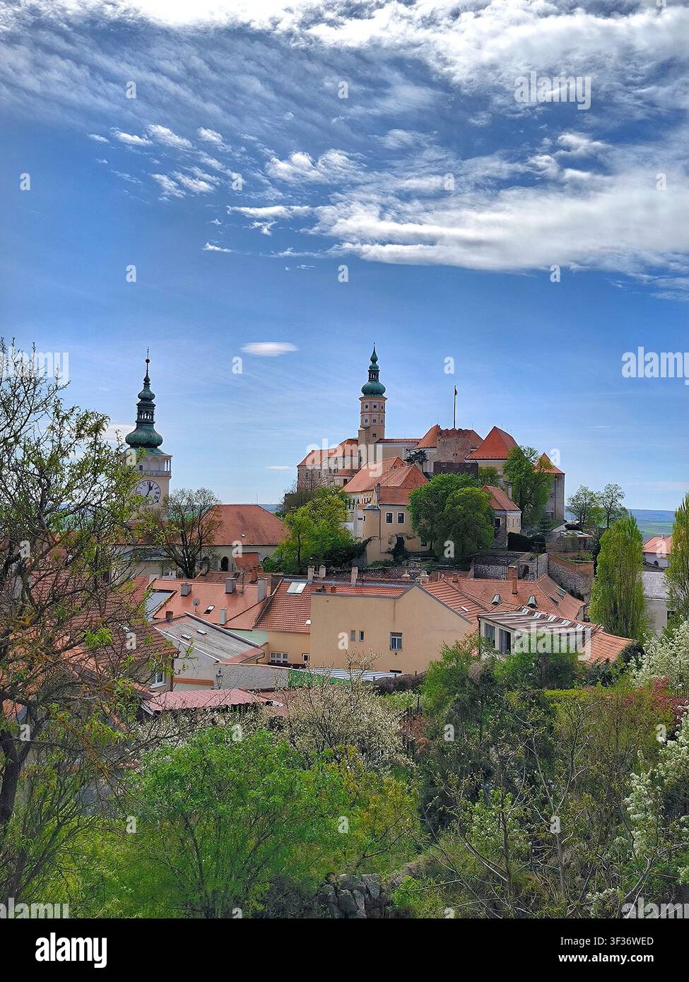 Blick auf das historische Schloss Mikulov und den Stadtturm, der sich über roten Dächern unter einem dramatischen blauen Himmel erhebt. - Smartphone-aufgenommenes Stockfoto