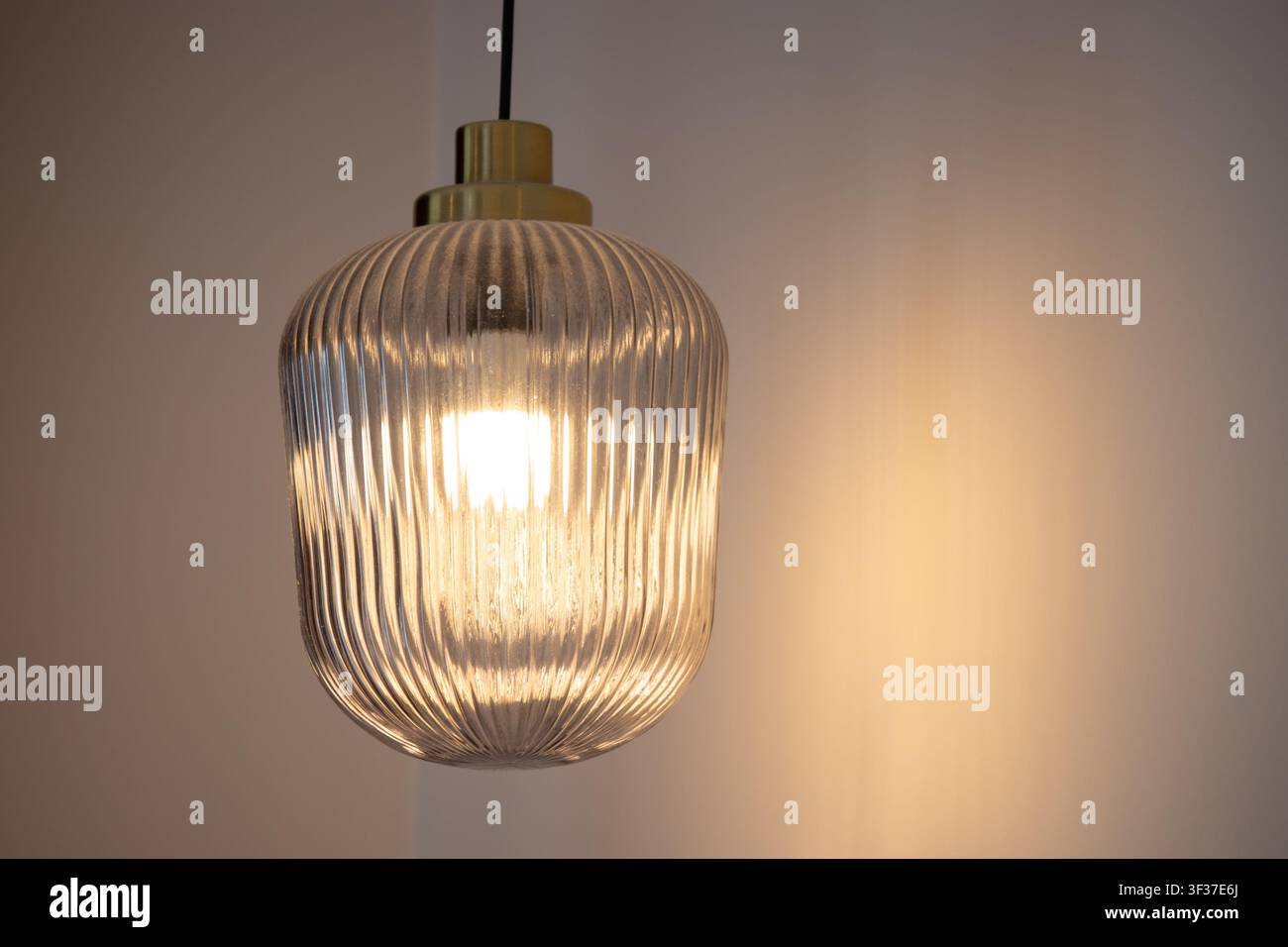 Nahaufnahme einer beleuchteten modernen Pendelleuchte mit geriffeltem Glasschirm und Messingarmatur, die ein warmes Licht in einem minimalistischen Innenraum wirft Stockfoto