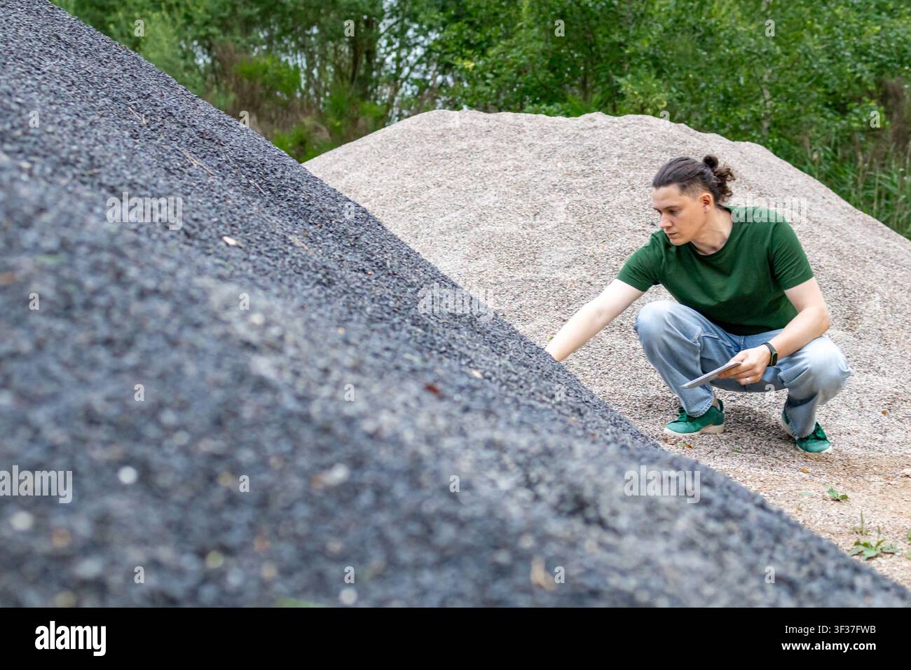 Mittealter Mann betrachtet einen Haufen dunklen Schotter, während er ein Tablet hält, während eines Besuchs vor Ort. Konzept der Materialbewertung, Ingenieurwesen Stockfoto