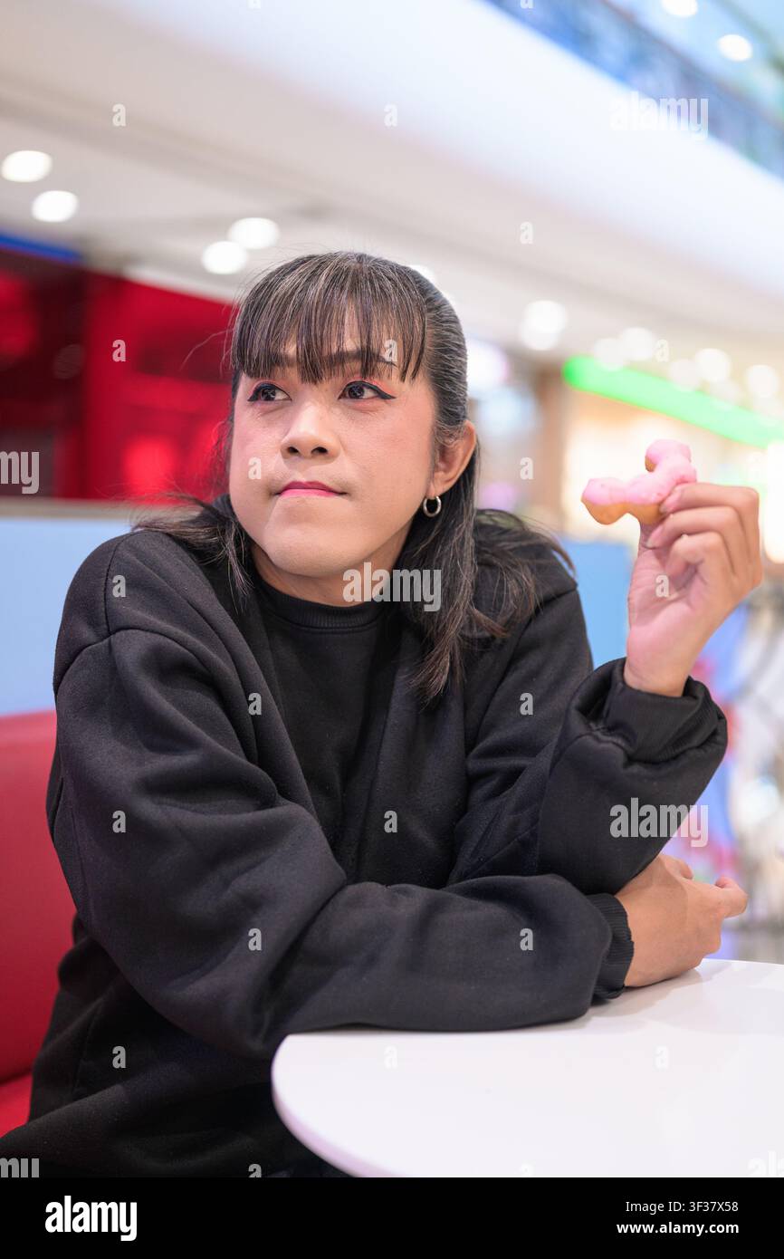 Asiatische Person, die einen Arm auf einem weißen Tisch abstützt und ein Stück eines rosa glasierten Donuts in die Luft hält, gedankenverloren zur Seite blickend mit einem Stockfoto