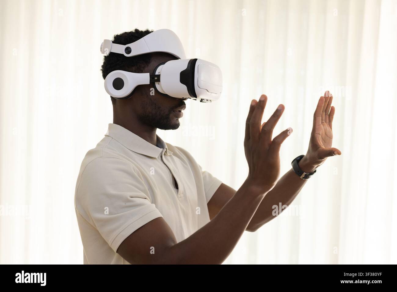 Afrikanischer Mann trägt ein beiges Polohemd, ein weißes VR-Headset, eine Smartwatch und gestikuliert mit den Händen in der Nähe durchsichtiger Vorhänge Stockfoto