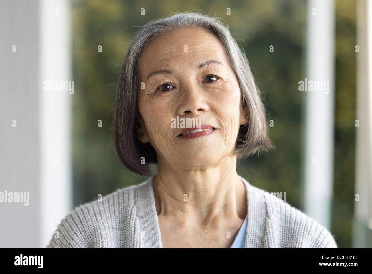 Senior asiatische Frau steht auf der Veranda und trägt eine graue Strickjacke, ein blaues Oberteil, Ohrstecker, Garten im Hintergrund Stockfoto