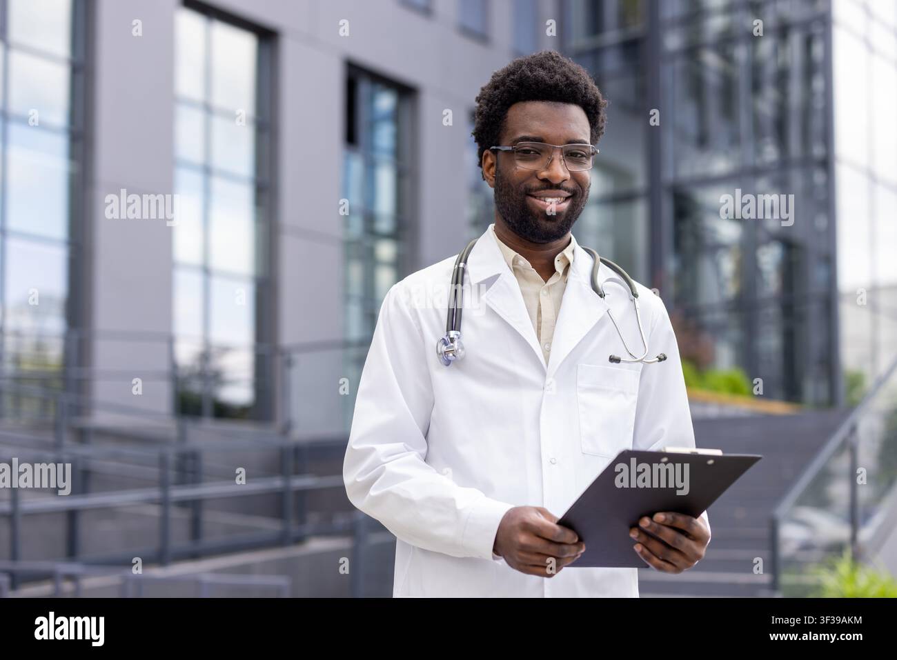 Junger afroamerikanischer Arzt trägt einen Laborkittel und ein Stethoskop. Selbstbewusst hält er ein Klemmbrett und lächelt in die Kamera vor einem modernen medizinischen Gebäude Stockfoto