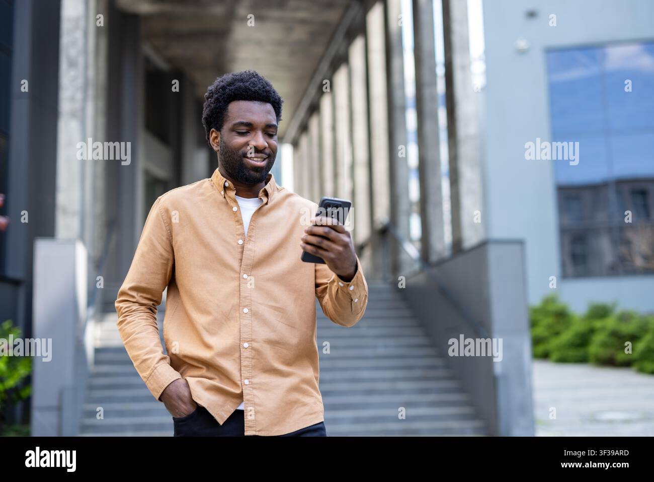 Junger Mann steht im Freien, lächelt, während er auf sein Mobilgerät schaut, verbindet und kommuniziert in einer städtischen Umgebung, spiegelt moderne Technologie und Lebensstil wider Stockfoto