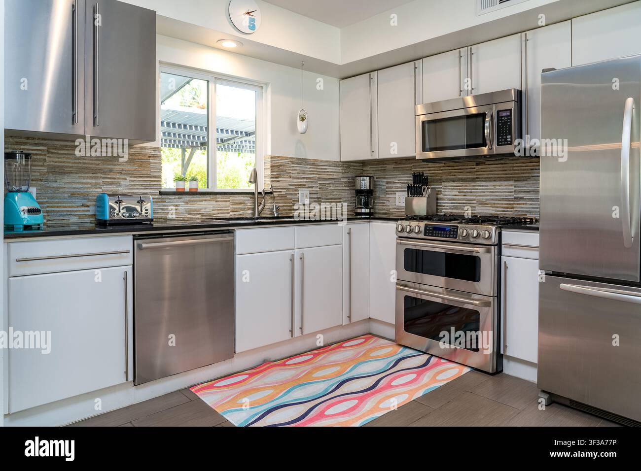 Modernes und helles Kücheninterieur. Edelstahlgeräte und weiße Schränke. Natürliches Licht, farbenfroher Teppich im Wohnraum. Stockfoto